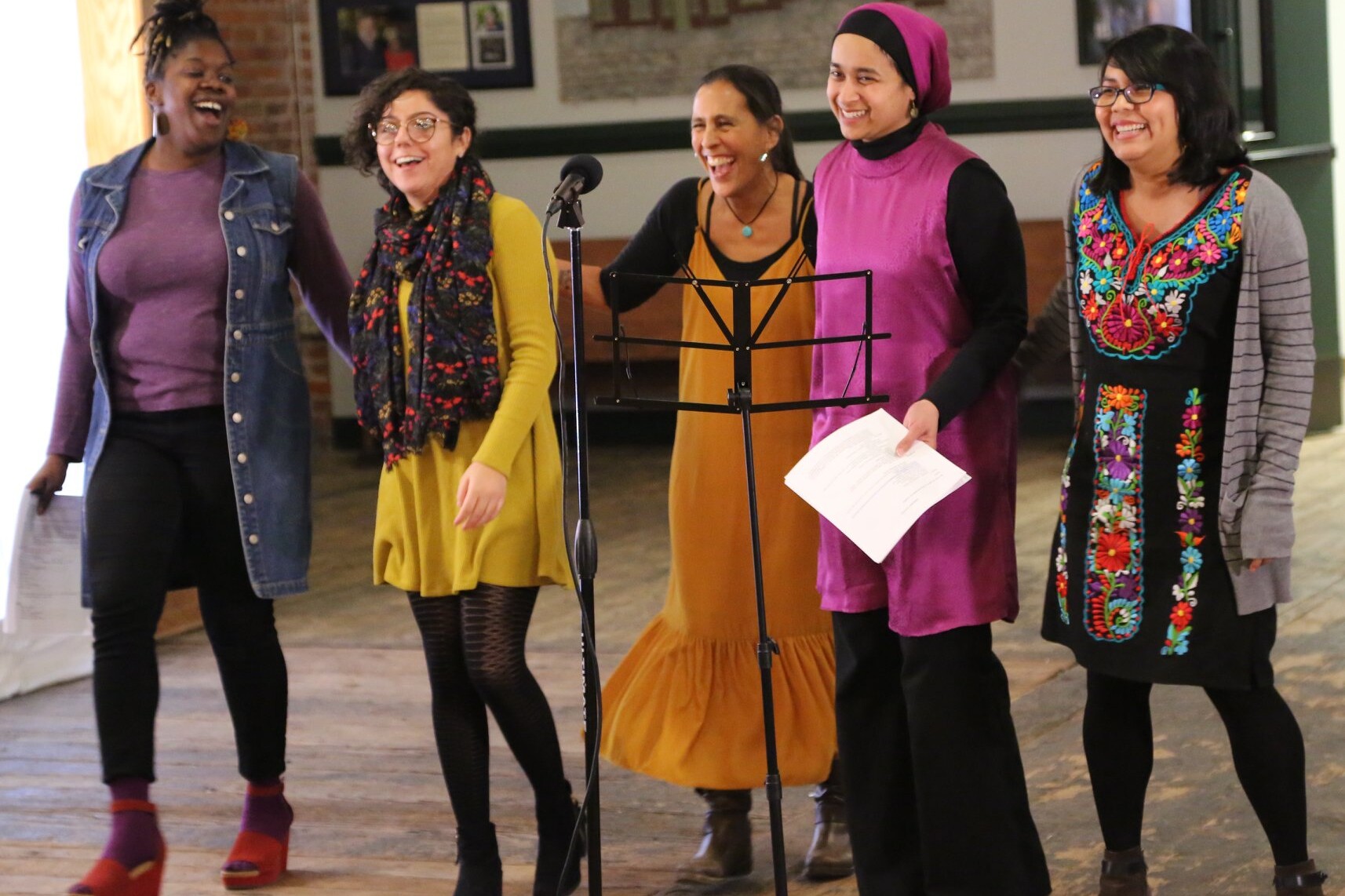 Five smiling and laughing BIPOC women stand in front of a microphone and music stand at New Moon Collaborations