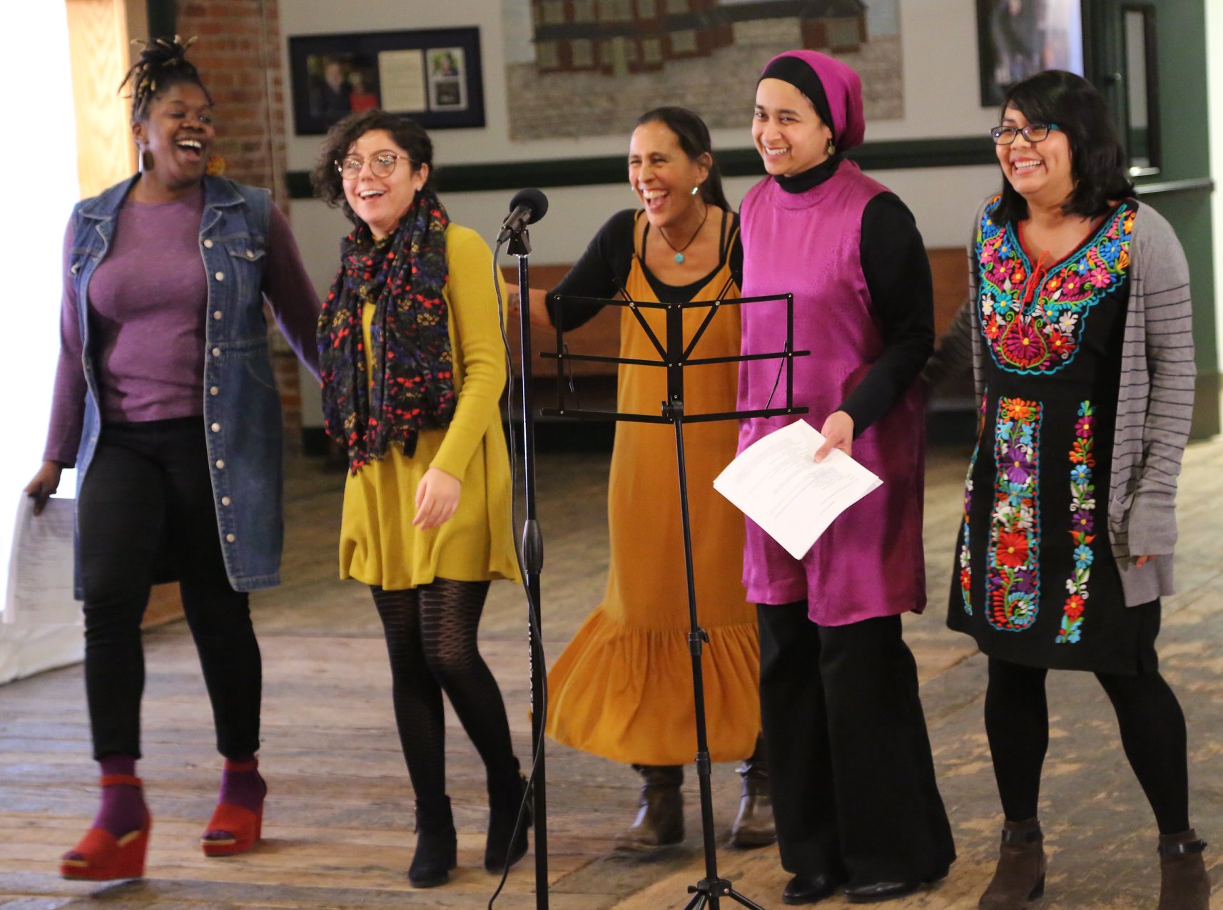 Five smiling and laughing BIPOC women stand in front of a microphone and music stand at New Moon Collaborations