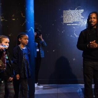 A black man talks to a woman and two children in a museum setting for Reimagine!