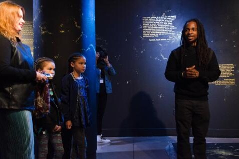 A black man talks to a woman and two children in a museum setting for Reimagine!