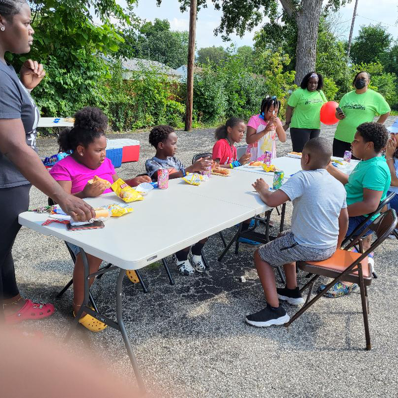 A group of seven children and four adults sure around a folding table eating snacks and socializing at Saginaw Just Transition Indaba