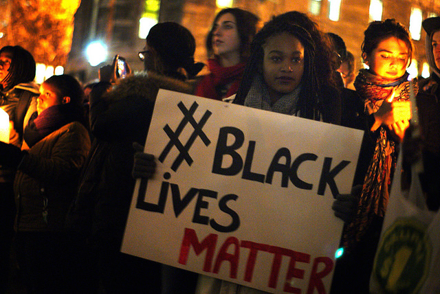 A person holds a poster board sign that reads #Black Lives Matter. They are surrounded by people holding candles in the dark of the night.