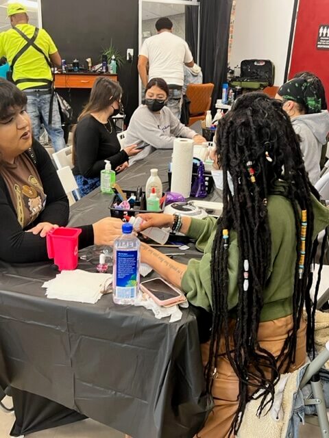 People sit around a table getting their nails done and socializing. Their are people getting their hair cut in the background.