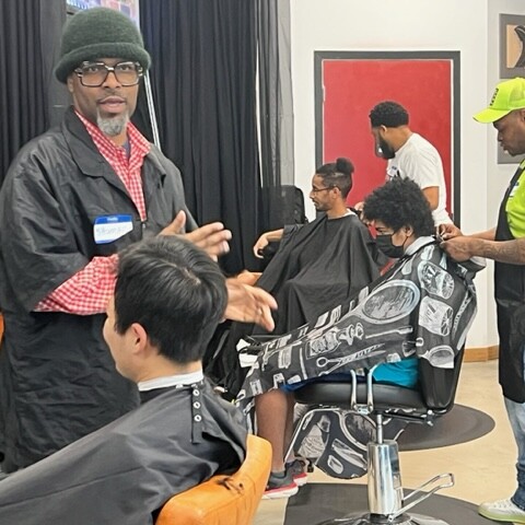 Two Black men cut to kids' hair with a third person and client tin the background.