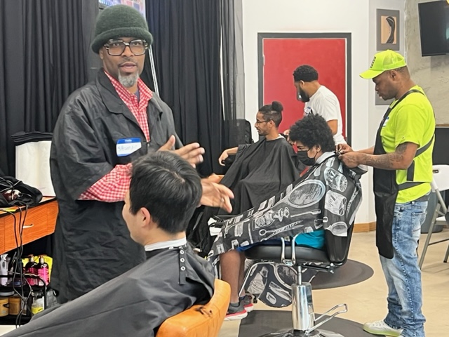Two Black men cut to kids' hair with a third person and client tin the background.