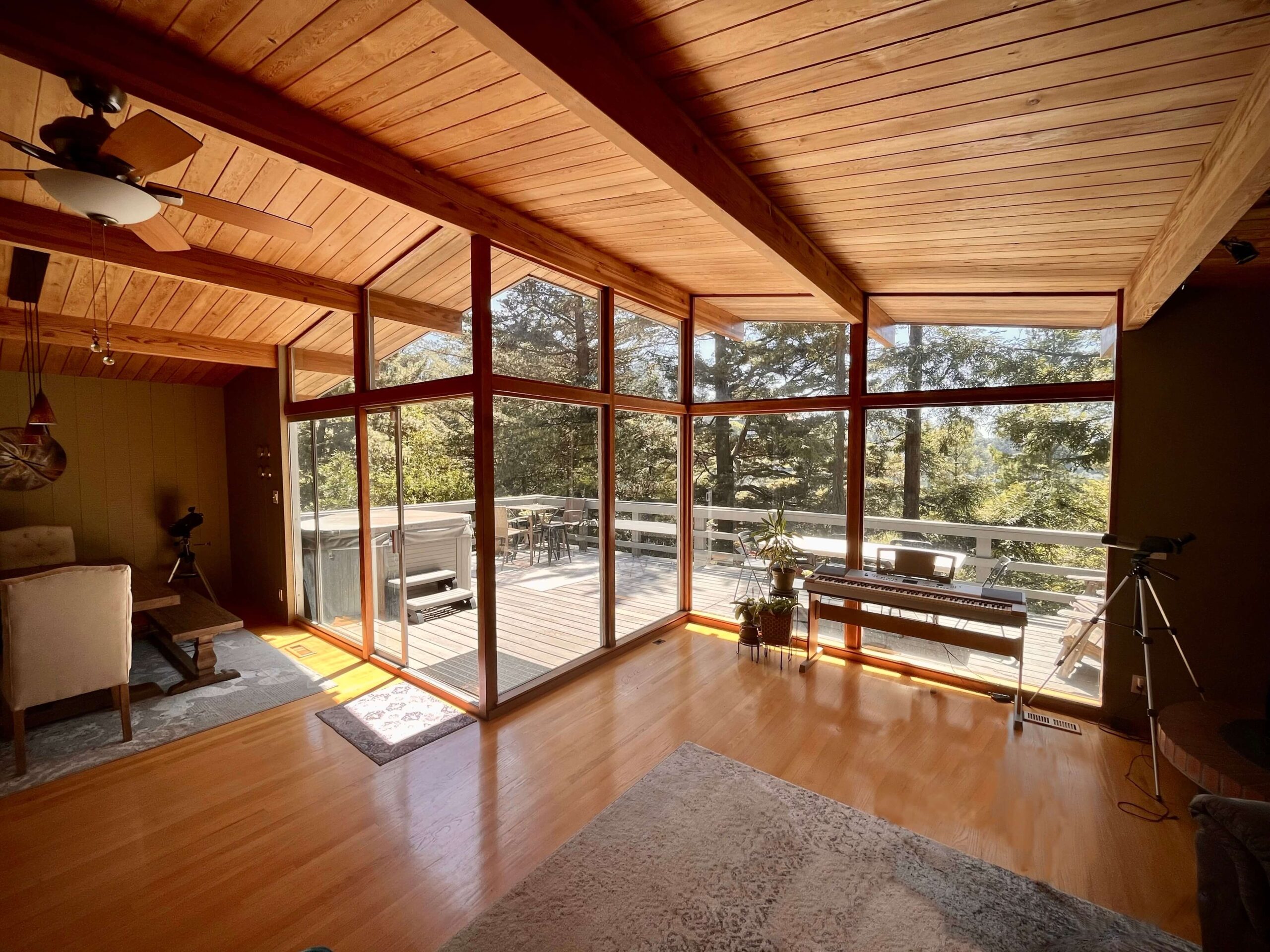 A large wooden interior roof open to a a wall of floor to ceiling glass windows. Outside is a wooden deck surrounded by the canopy of pine trees.
