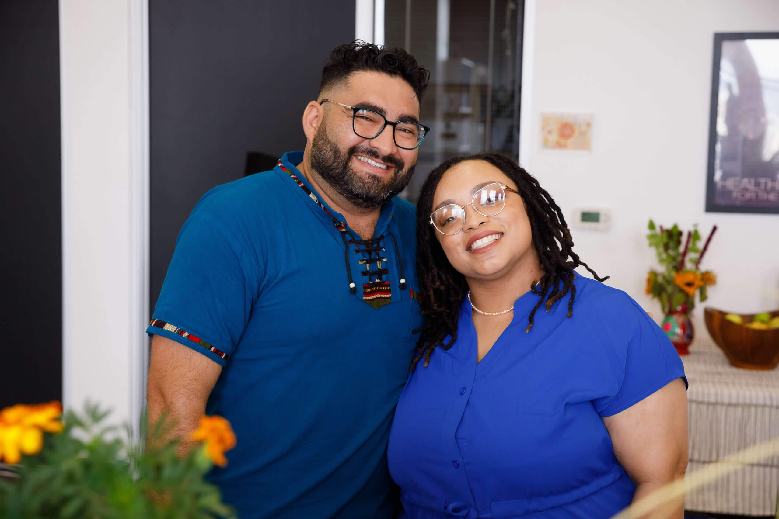 Two people both wearing blue shirts lean in together while smiling at the camera