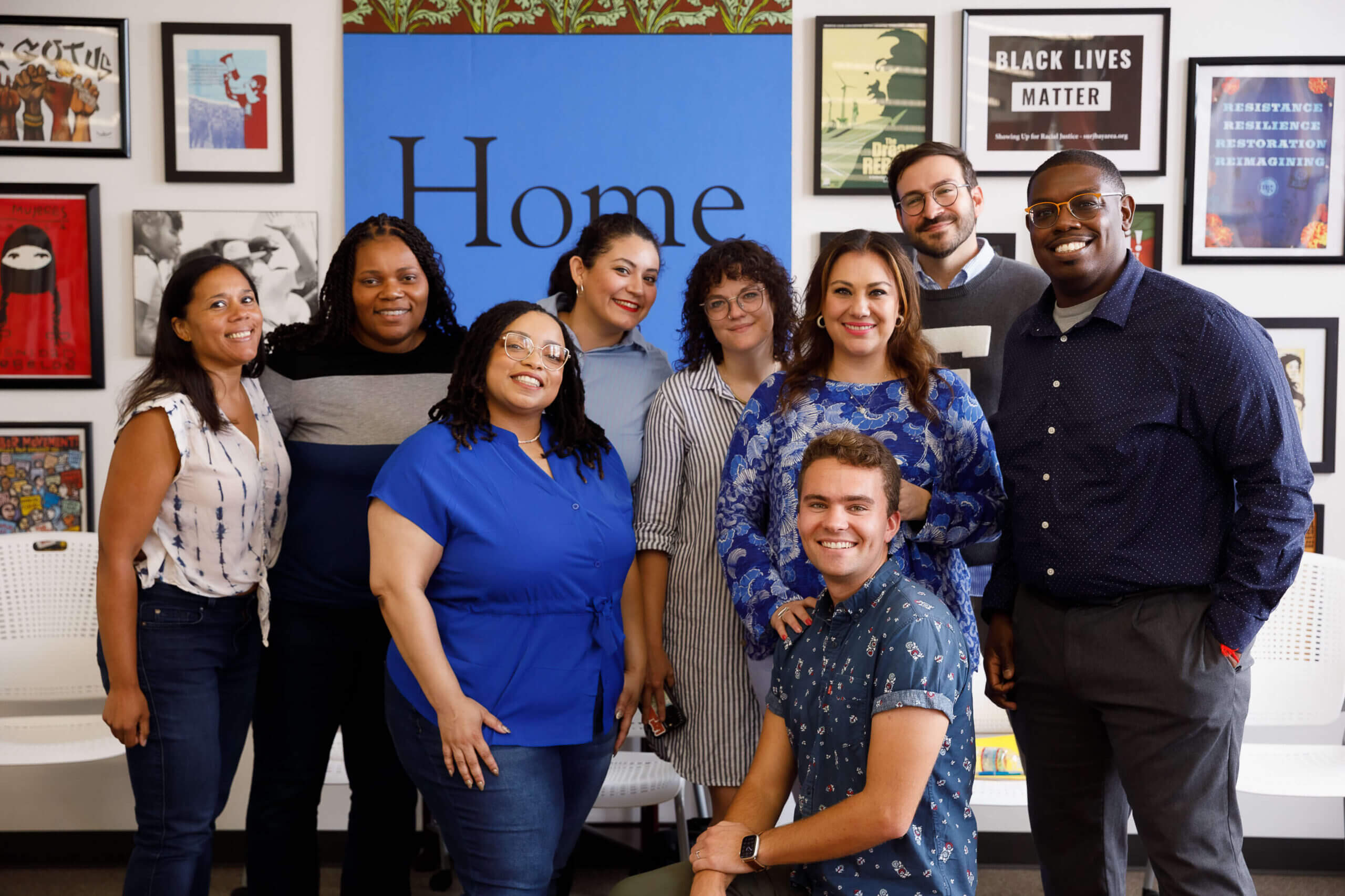 Nine people pose for a group photo while wearing blue shirts. Most are standing and one is kneeling in front. They are in front of a wall full of framed posters and art.