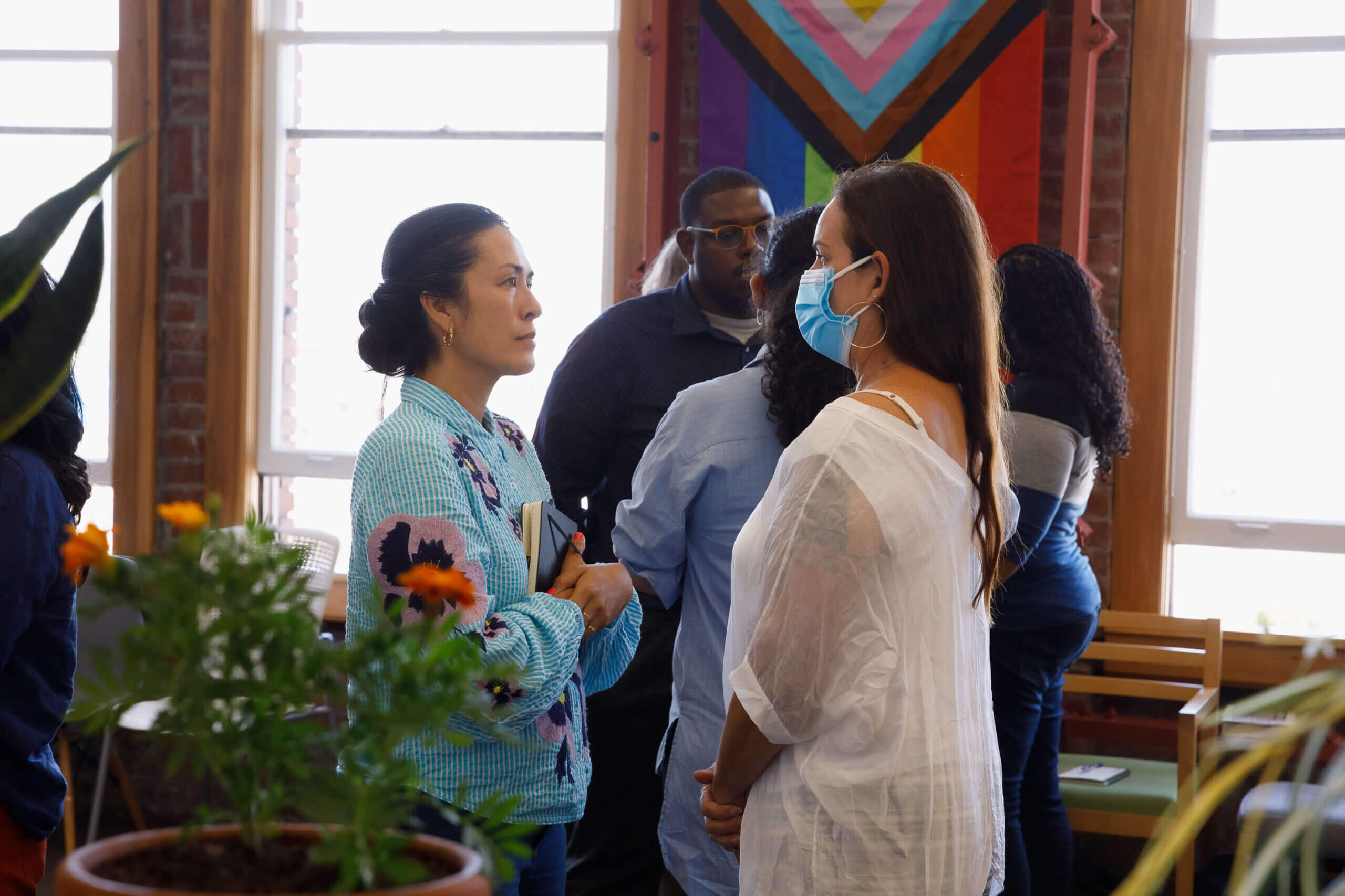 Two people talk, one is wearing a mask as she talks. There are groups of other people talking with a pride flag hanging in the background.