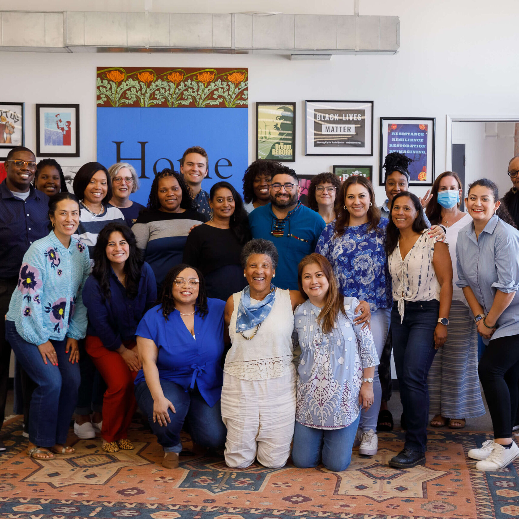 Twenty three people wearing blue shirts smile for a group photo in three separate rows. A wall full of framed posters and activist art is behind them.