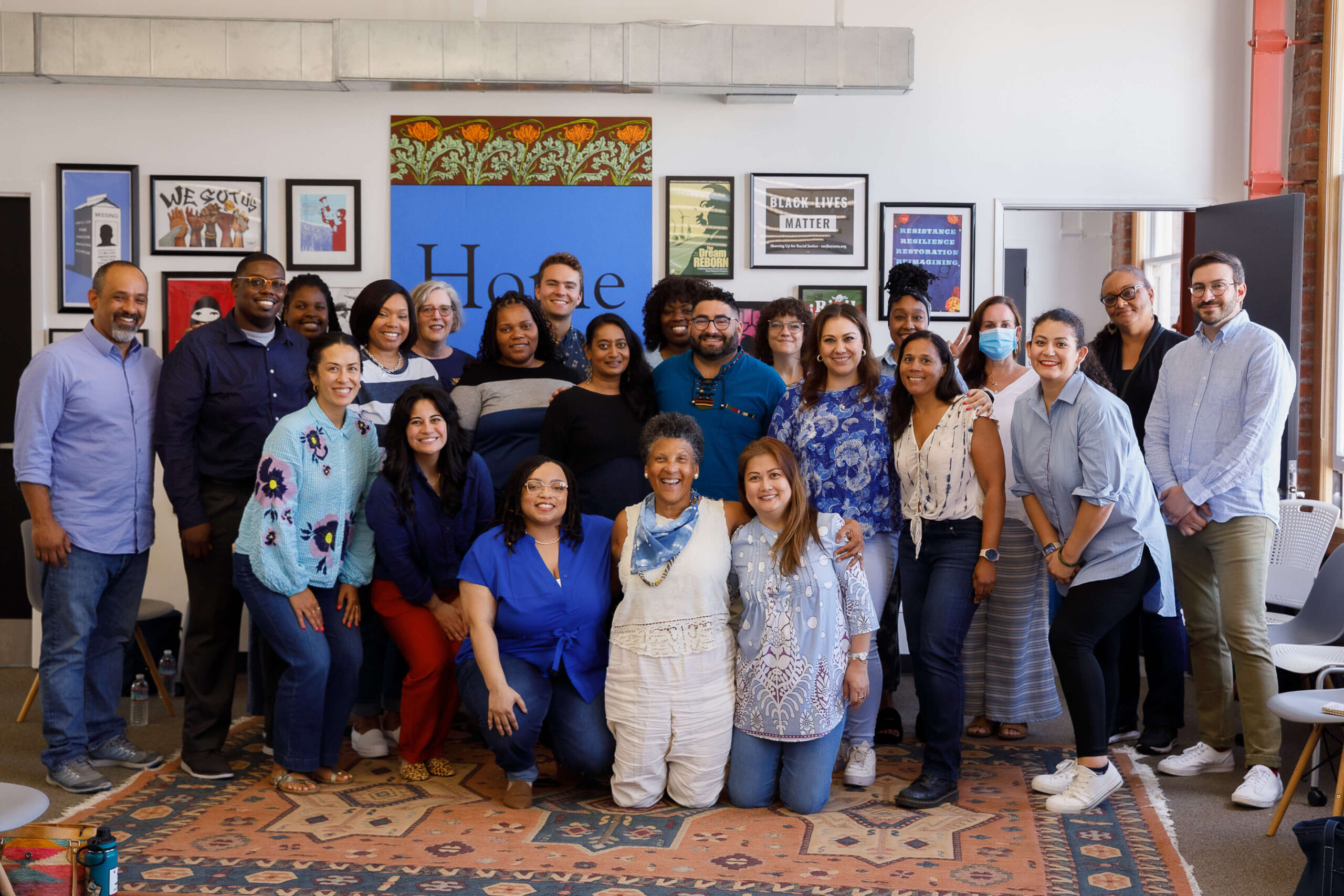 Twenty three people wearing blue shirts smile for a group photo in three separate rows. A wall full of framed posters and activist art is behind them.